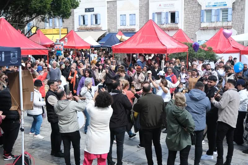 Meydanda halka olmuş kalabalık; ortada davul çalınıyor, çevrede kırmızı çadırlar ve dükkânlar