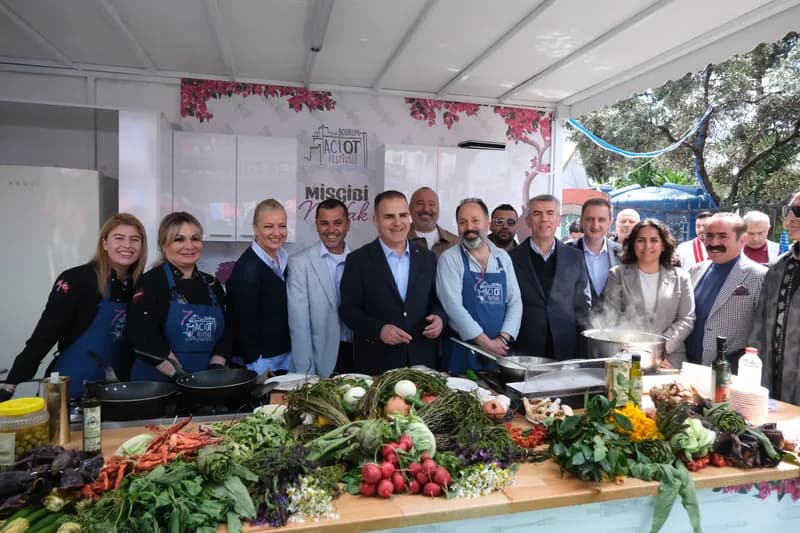 Festival önlüklü ekip, önlerinde turp, enginar, taze ot demetleri ve zeytinyağı şişeleriyle dolu uzun tezgâhın arkasında